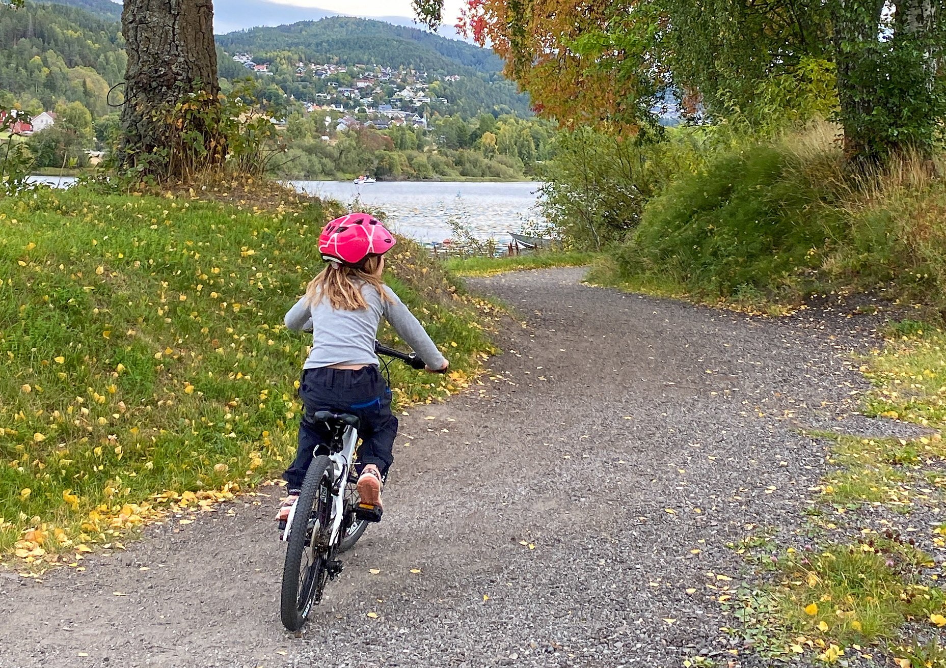 En jente bakfra, med rosa sykkelhjelm, sykler på en sti ved et vann