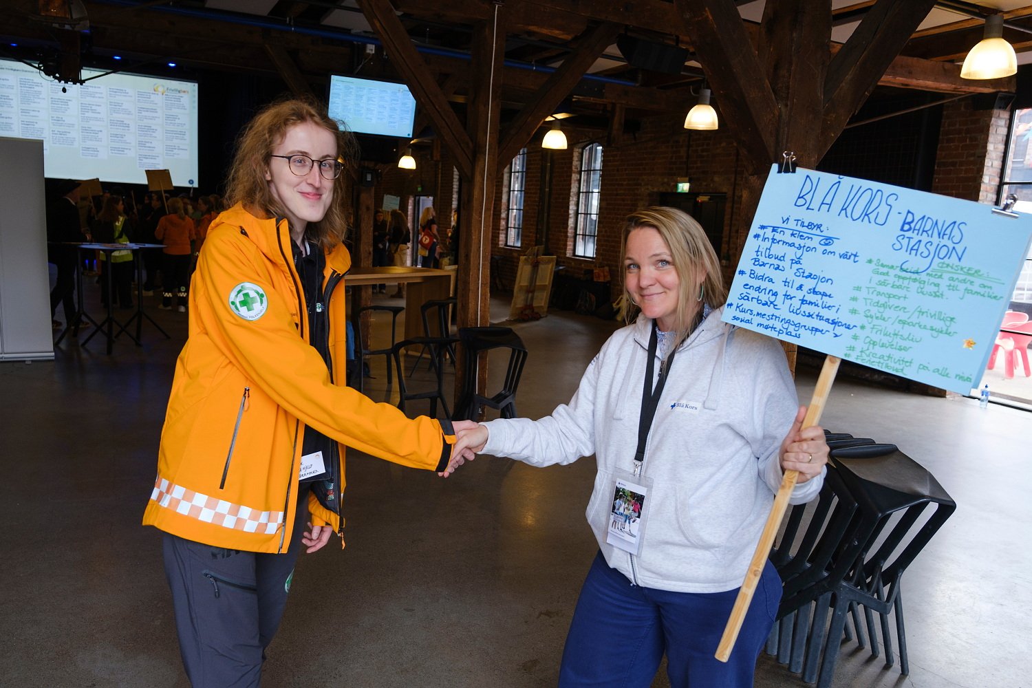 To kvinner tar hverandre i hånda og smiler til kamera. Kvinnen til venstre har en oransje jakke med reflekser og kvinnen til høyre har en blå plakat med mye tekst på i hånda.