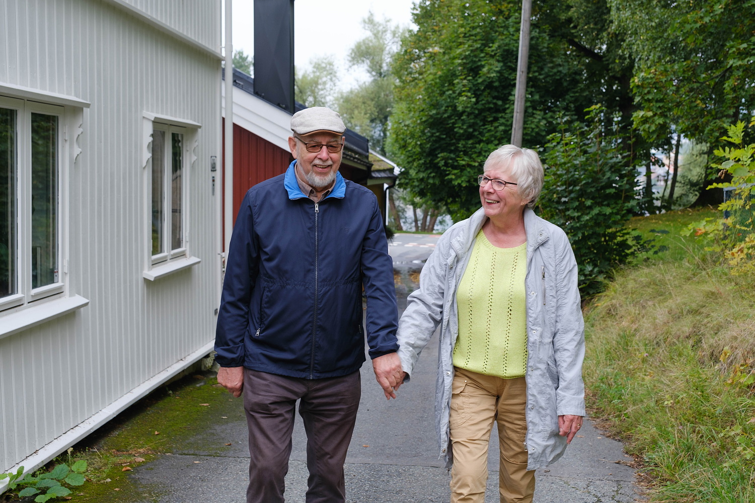 Et eldre par går tur på en asfaltert gangsti. Ved siden av er det et par små hus i trebebyggelse. De smiler.