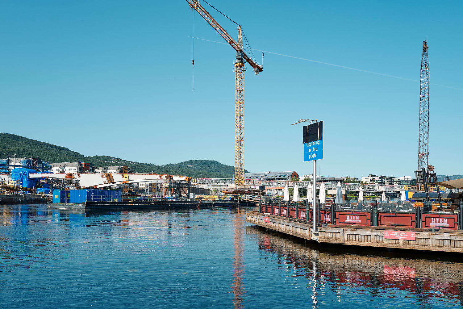 Elv med anleggsarbeider og heisekran og en brygge med restaurant til høyre i bildet.