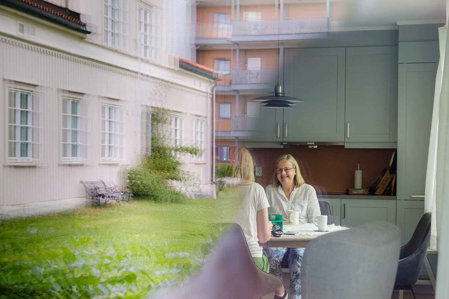 To damer som snakker sammen, ansiktet synes kun på hun eldre. De sitter ved et kjøkkenbord i en leilighet. I vinduene reflekteres også et trehus.