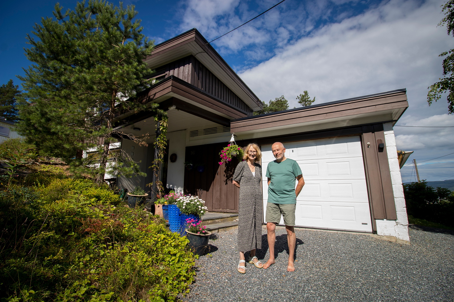 Ivar Flaten og Ingvil Tågvold Flaten står foran huset sitt. Det er sol, det brune huset bak dem er deres. 