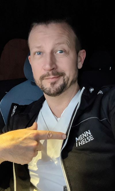 Mann peker på jakka si mens han sitter i bilen. På jakka står det "Menn  helse".