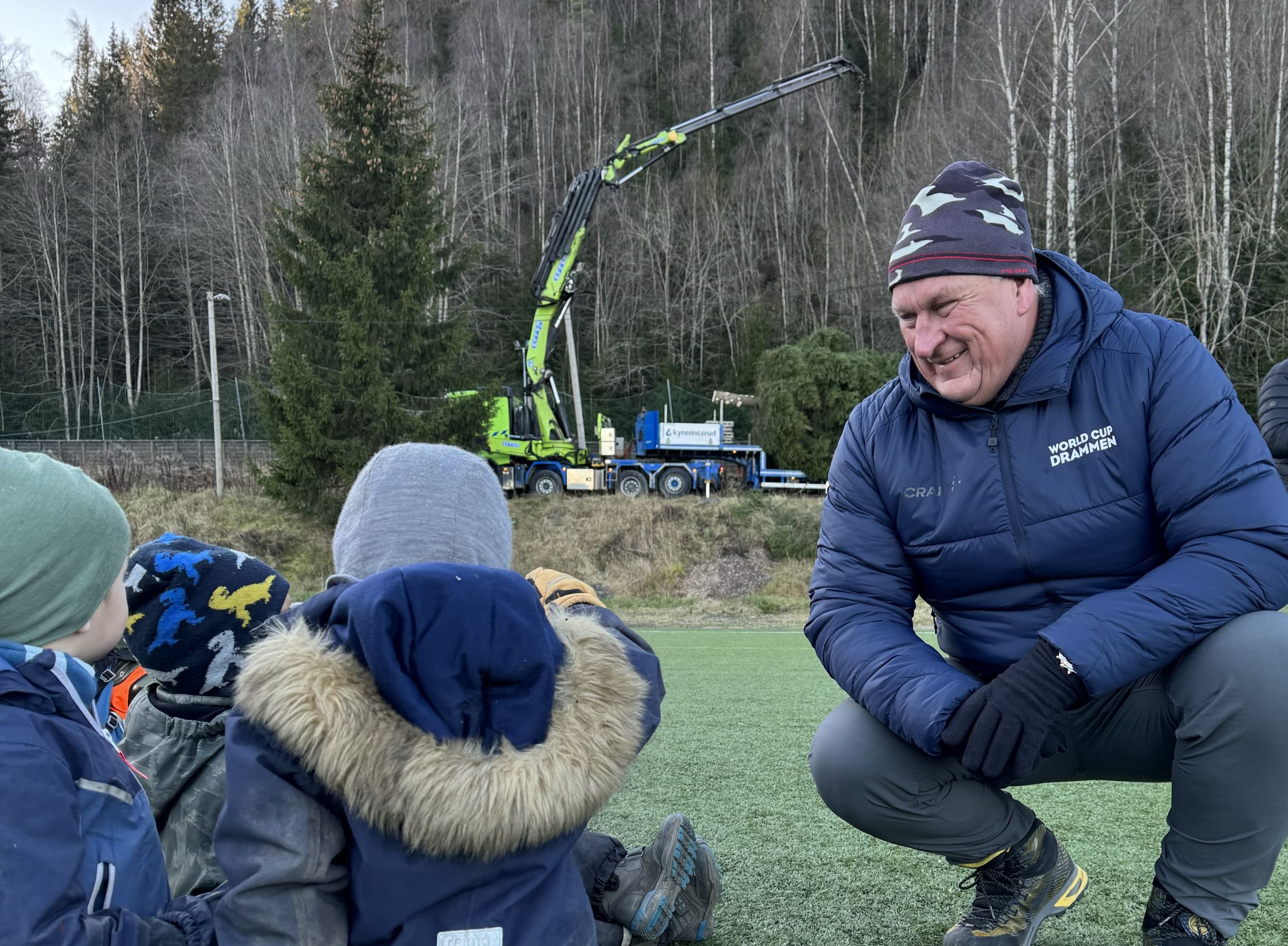 en mann prater med barn. I bakgrunnen et stort grantre som legges på et lasteplan.