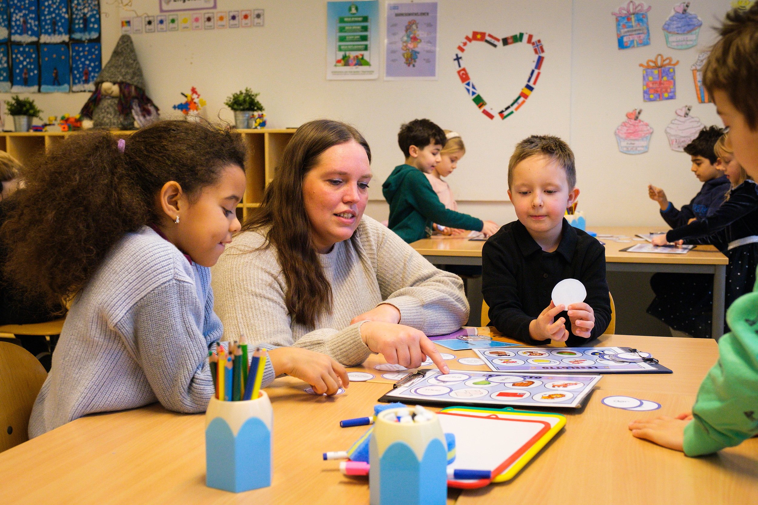 En kvinnelig lærer viser en gutt og en jente en oppgave. De sitter ved en skolepult. 